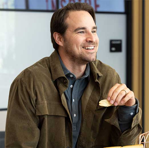 A man with brown hair and a beard, wearing a brown jacket and grey shirt, smiles while holding a chip from his meal at a sandwich franchise. He is sitting indoors.