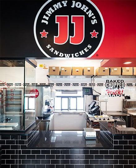 Interior of a Jimmy John's sandwich shop showing the counter, cash register, stacked supplies, and vibrant decor representing the sandwich franchise.