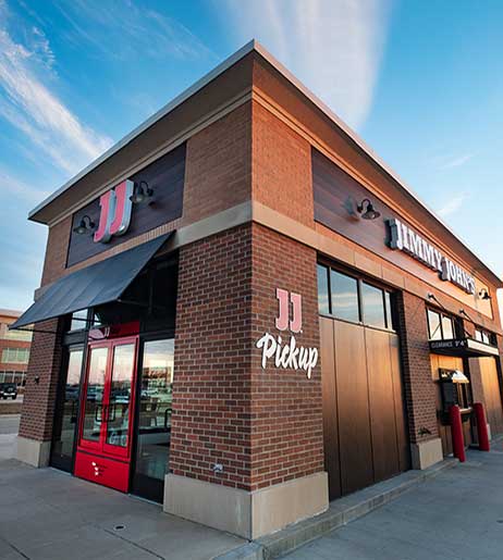 Exterior view of a Jimmy John's sandwich franchise at dusk, featuring a brick building with prominent signage and a red door.