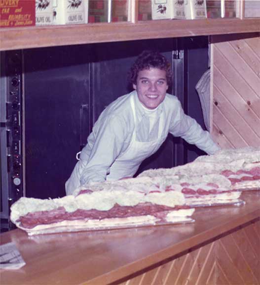 Young man in a white uniform preparing a large sandwich at a sandwich franchise counter, smiling at the camera.