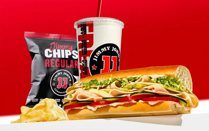 A jimmy john's sub sandwich from the sandwich franchise with lettuce, tomatoes, and meats, accompanied by a bag of jimmy chips and a soda cup.